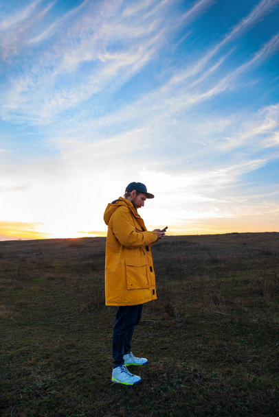 Traveler άνθρωπος σε κίτρινο παλτό με smartphone με τα πόδια σε ένα λόφο ηλιοβασίλεμα θέα στον ουρανό πλευρά. Αυθεντικό αρσενικό τουρίστα με τρόπο ζωής gadget. Hipster τύπος υπαίθρια Μεμονωμένα ταξίδια περιπέτεια. Ενεργό περπάτημα σακίδιο. - Φωτογραφία, εικόνα