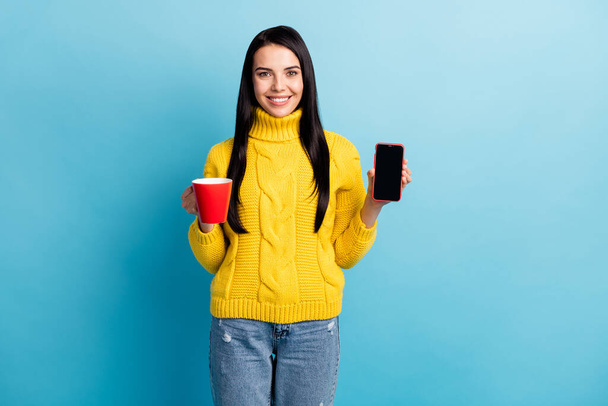 Photo portrait of woman smiling keeping mug with beverage showing cellphone screen with copyspace isolated on vibrant blue color background. - Photo, image