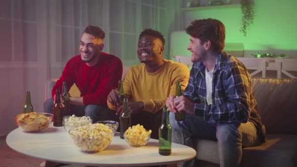 Trzech wielonarodowych facetów cieszących się razem piątkowym wieczorem, oglądających telewizję i wiwatujących z piwem w domu w neonowych światłach - Materiał filmowy, wideo