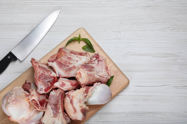 Cutting board with raw chopped meaty bones, basil and knife on white wooden table, flat lay. Space for text - Фото, зображення