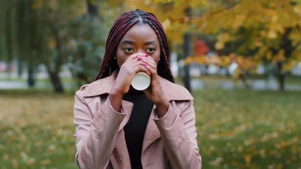 Мечтательная афроамериканская молодая женщина пьет кофе или чай в одноразовой чашке, задумчивая студентка тысячелетия наслаждается ароматным горячим напитком в пасмурный осенний день. У работницы перерыв на открытом воздухе - Кадры, видео