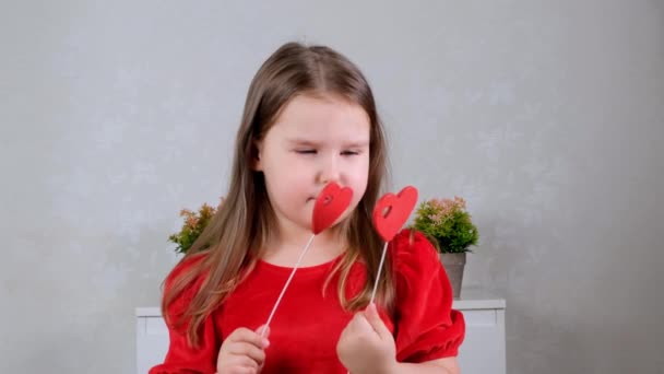 grappig mooi klein meisje in een rode jurk met twee harten op een stok sluit haar ogen met hen. Valentijnsdag concept. - Video