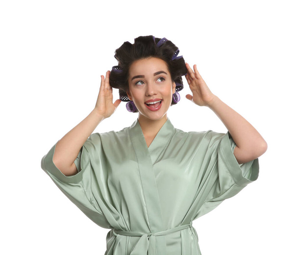 Happy young woman in silk bathrobe with hair curlers on white background - Fotoğraf, Görsel