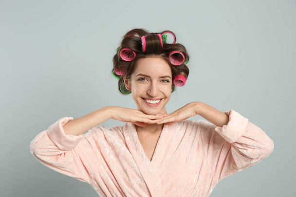 Beautiful young woman in bathrobe with hair curlers on light grey background - Foto, Imagen