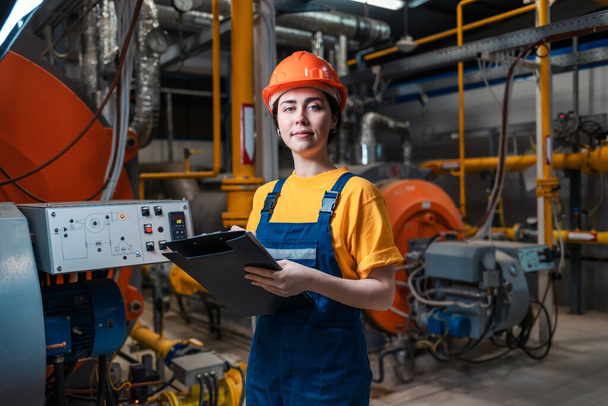 Concept van een industriële fabriek. Een glimlachende vrouw in uniform en helm met een map en een pen in haar hand. In de achtergrond-stookruimte. - Foto, afbeelding