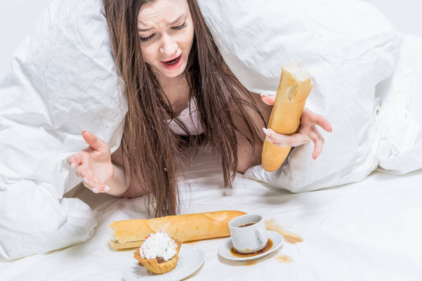 Breakfast. The girl is indignant, spilled coffee on the bed. The brunette lies in bed, next to her is a cupcake, a baguette in her hand and a cup. - Foto, Imagen