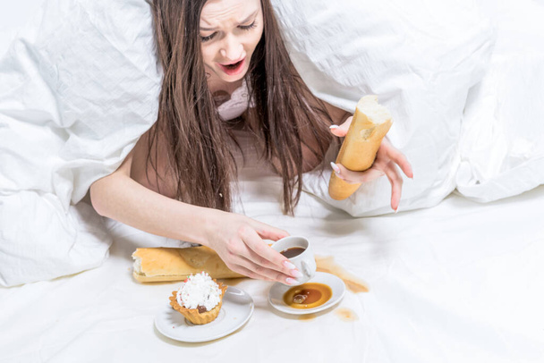 The girl is indignant, spilled coffee on the bed. Breakfast. The brunette lies in bed, next to her is a cupcake, a baguette in her hand and a cup. - Photo, Image