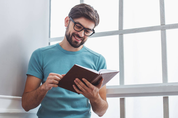 Bonito homem em roupas casuais e óculos lendo um livro e sorrindo enquanto sentado perto da janela
 - Foto, Imagem