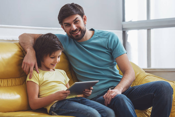 Handsome young father in casual clothes looking in camera and smiling while sitting on a sofa near his cute little son, who using a tablet - Photo, Image