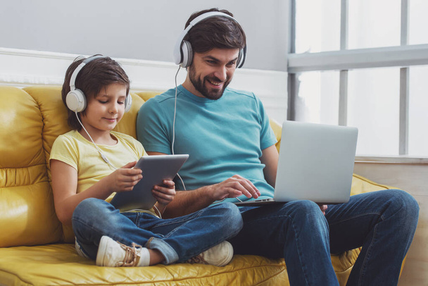 Handsome young father in casual clothes using a laptop and his cute little son using a tablet while sitting on a sofa in the room. Both in headphones. - 写真・画像