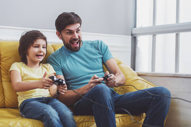 Bonito pai jovem em roupas casuais e seu filho pequeno bonito jogando jogos de consola enquanto sentado em um sofá na sala
 - Foto, Imagem
