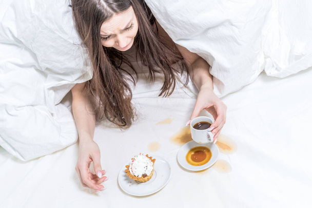 The girl is indignant, spilled coffee on the bed. The brunette lies in bed, next to her is a cupcake, a baguette in her hand and a cup. Breakfast. - Foto, Imagen