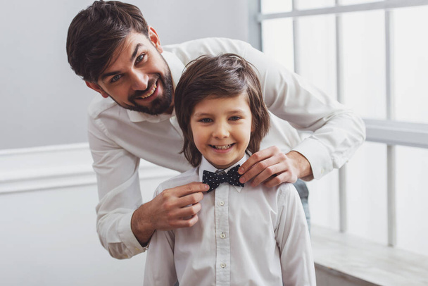 Handsome young father adjusting his cute little son's bow tie. Both white classical shirts looking in camera and smiling - Fotó, kép