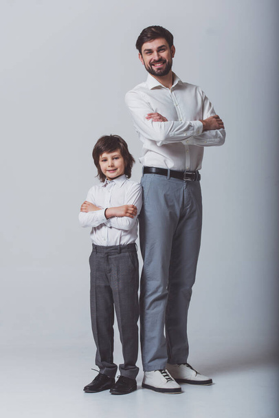 Portrait of a handsome young father and his cute little son standing back to back, looking in camera and smiling. Both in white classical shirts and gray pants, standing on a gray background. - Fotó, kép