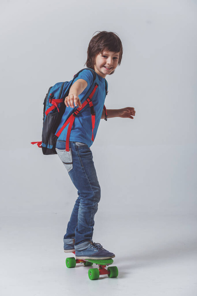 Portret van een schattige kleine schooljongen in een blauw t-shirt met een rugzak in de camera kijken en glimlachen terwijl het skateboarden op een grijze achtergrond - Foto, afbeelding