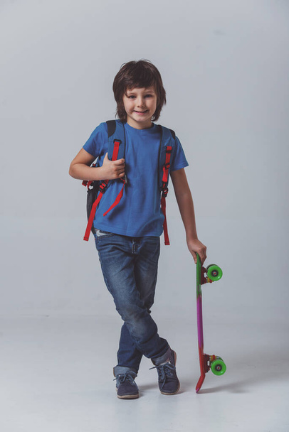 Cute little schoolboy in a blue t-shirt with a backpack holding a skateboard, looking in camera and smiling while standing on a gray background - Fotografie, Obrázek