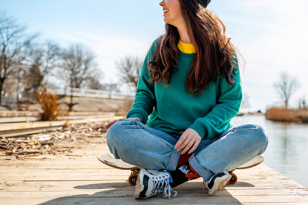 A young Caucasian woman sits cross-legged on a skateboard and looks away with a smile. In the background-the sea and the coastline. Close up. Concept of sports lifestyle and street culture. - Fotó, kép