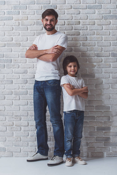Красивий молодий батько і його милий маленький син, що стоїть спиною, дивлячись в камеру і посміхаючись. І в білих футболках, і в джинсах, що стоять на стіні з білої цегли
. - Фото, зображення