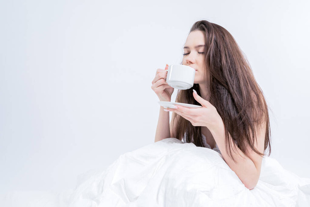 A beautiful woman is sitting in bed and drinking aromatic coffee. Tasty breakfast. The brunette is holding a cup with a hot drink. White bedroom. Good morning. - Fotoğraf, Görsel