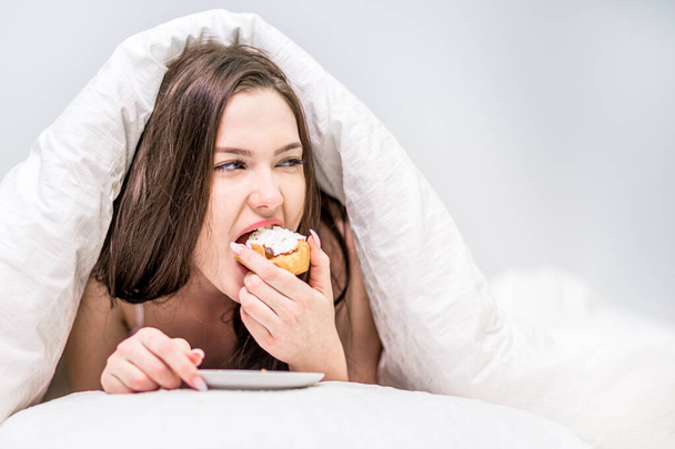 Calories. A cute woman with appetite eats a cupcake lying on the bed under the covers. Dessert. Breakfast in bed. - Фото, изображение