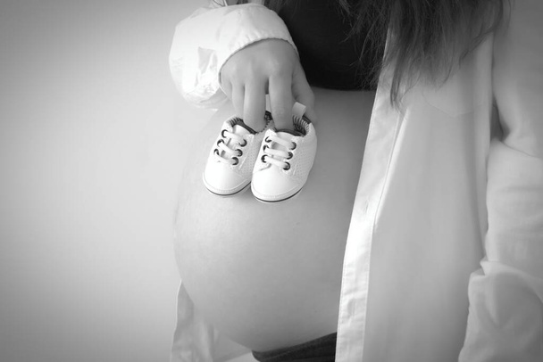 Uma mulher em seu 8o mês de gravidez mostra sua barriga e segura minúsculos sapatos de bebê.Conceito cinza - barriga de gravidez na semana 32 e sapatos de bebê. - Foto, Imagem