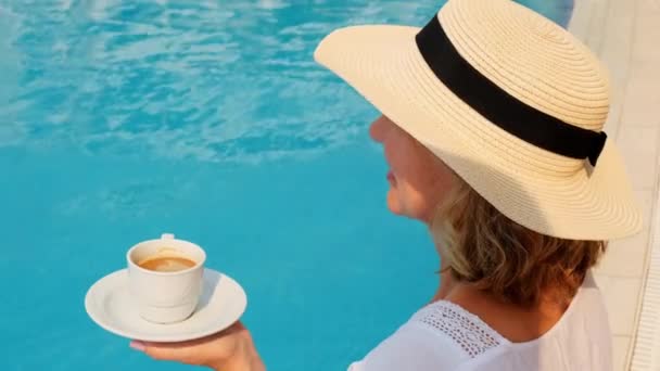 50-jährige Frau mit Strohhut im weißen Kleid entspannt am Pool bei einer Tasse Kaffee, Urlaubskonzept, guten Morgen. Frau sitzt am Pool. - Filmmaterial, Video