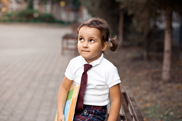 Μικρή χαριτωμένη μαθήτρια με στολή να κάθεται σε ένα παγκάκι και να γελάει - Φωτογραφία, εικόνα