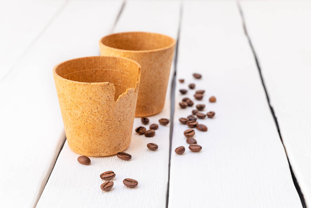 Šetrné k životnímu prostředí vafle šálky kávy s praženými kávovými zrny na bílém dřevěném pozadí - Fotografie, Obrázek