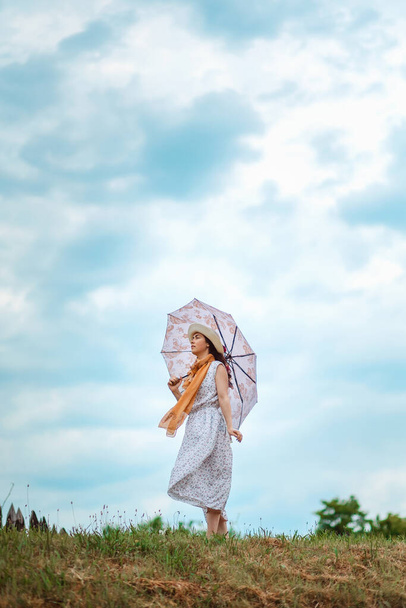 Nuori nainen olkihatussa, mekossa, sateenvarjo käsissään seisoo kukkulalla ja katsoo pilvistä taivasta. Kopioi tilaa. Psykologian käsite. - Valokuva, kuva