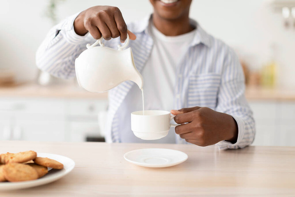 Satisfied young black woman pours milk at cup of favorite drink, sits at table with cookies in kitchen interior - Fotó, kép