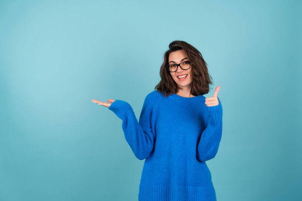 Une femme en pull bleu tricoté, cheveux bouclés courts, lunettes sur les yeux, sourit joyeusement, pointe vers un espace vide, tient dans sa paume - Photo, image
