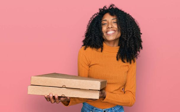 Mladá africká americká dívka drží dodávku pizza box vypadá pozitivně a šťastně stojící a usmívá se s sebevědomým úsměvem ukazující zuby  - Fotografie, Obrázek
