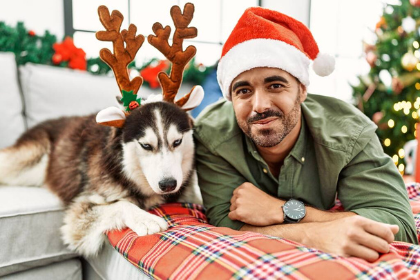 Young hispanic man lying on sofa with dog wearing reindeer hat by christmas tree at home - Fotoğraf, Görsel