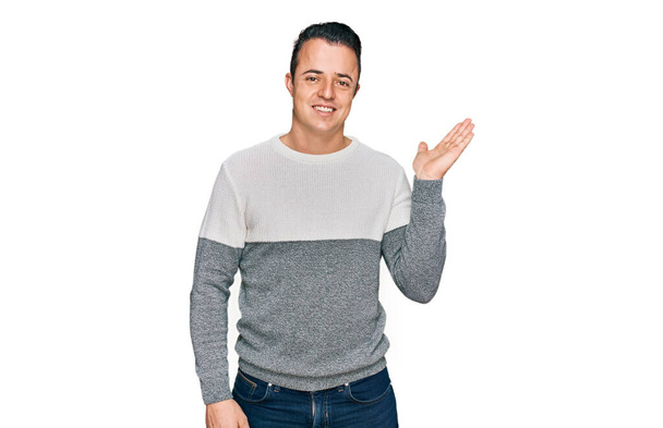 Hezký mladý muž na sobě ležérní zimní svetr s úsměvem veselý prezentaci a ukazuje s dlaní ruky při pohledu do kamery.  - Fotografie, Obrázek