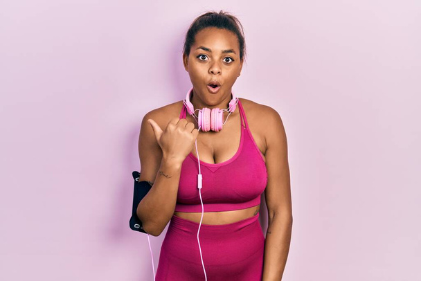 Молода афроамериканка, одягнена в спортивний одяг і з навушниками, здивований вказуючи пальцем вбік, з відкритим ротом..  - Фото, зображення