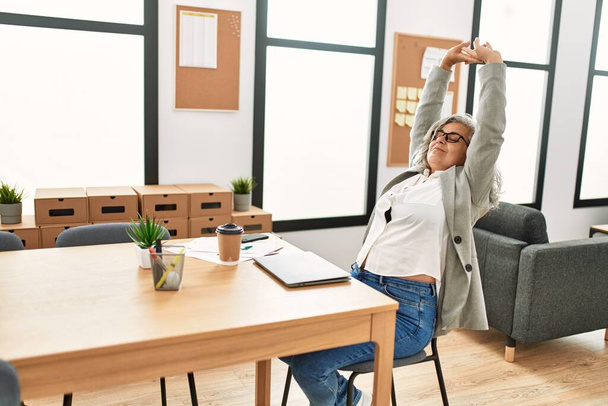 Middle age grey-haired businesswoman tired stretching arms working at the office. - Φωτογραφία, εικόνα