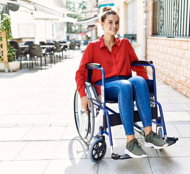 Jonge vrouw glimlachend zelfverzekerd zittend op rolstoel op straat - Foto, afbeelding