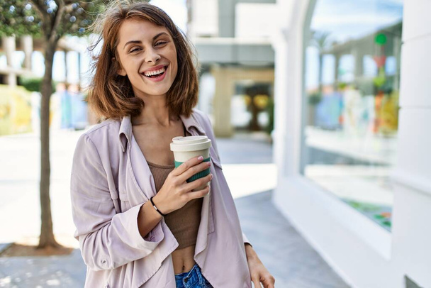Joven chica caucásica sonriendo feliz bebiendo café en la ciudad. - Foto, imagen