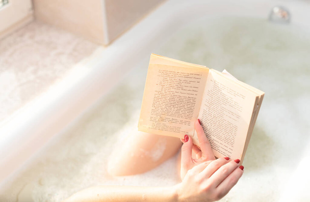 Young woman takes a bath with lush foam near the window reading the book close up. Personal hygiene, healthcare, relax at home concept. Selective focus - Foto, Imagen