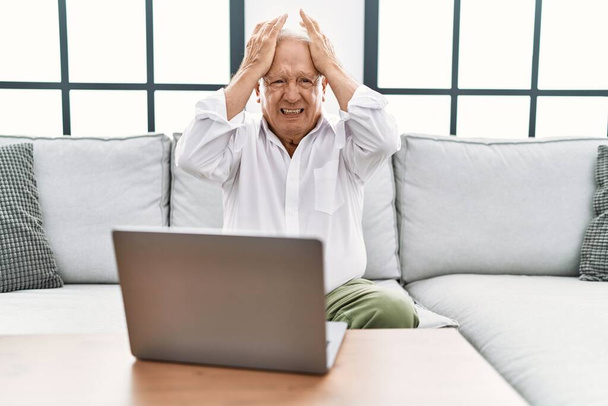 Старший использует ноутбук дома, сидя на диване, страдая от головной боли, отчаяния и стресса из-за боли и мигрени. руки на голове.  - Фото, изображение