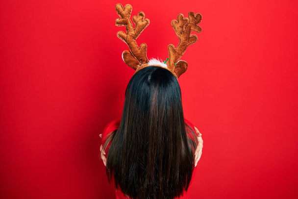 Молодая латиноамериканка в рождественской шляпе оленя и красном носу, обнимающая себя счастливой и позитивной с обратной стороны. любовь к себе и забота о себе  - Фото, изображение