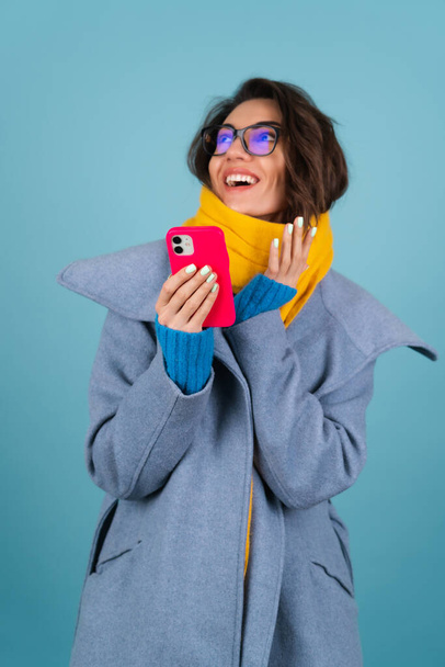 Spring autumn portrait of a woman in a blue knitted sweater, glasses, a yellow bright scarf and a gray coat, holds a phone and laughs merrily - Photo, Image