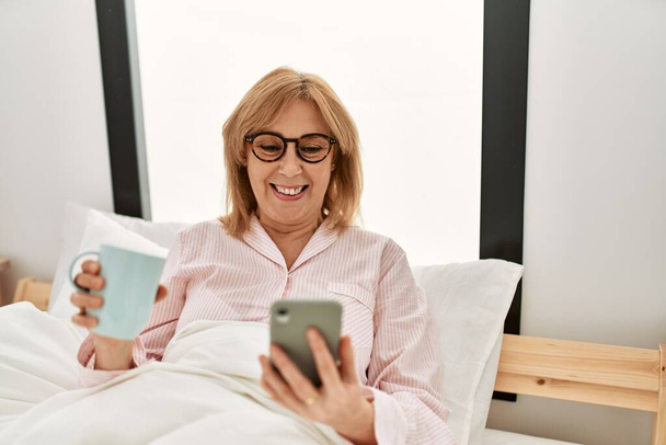 Donna bionda di mezza età sdraiata sul letto usando smartphone e bevendo caffè a casa. - Foto, immagini