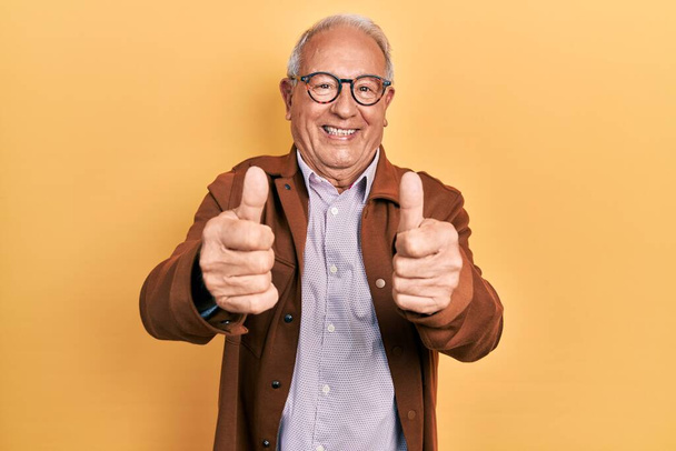 Starší muž s šedivými vlasy v neformální bundě a brýlích schvalující dělá pozitivní gesto rukou, palce nahoru s úsměvem a šťastný za úspěch. gesto vítěze.  - Fotografie, Obrázek