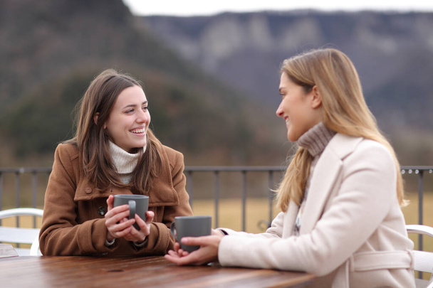 Two women drinking coffee and talking in winter sitting in a terrace - Фото, изображение