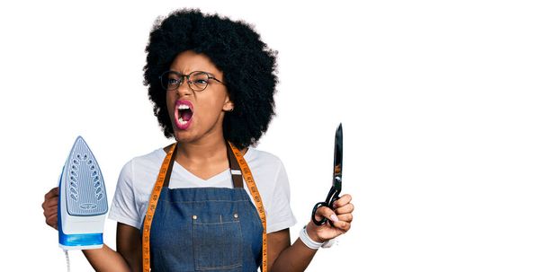 Junge afrikanisch-amerikanische Schneiderin Designerin in Atelierschürze hält Eisen und Schere wütend und wütend schreien frustriert und wütend, schreit vor Wut. Wut und aggressives Konzept.  - Foto, Bild