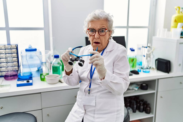 Idősebb nő ősz hajjal dolgozik tudós laboratóriumban nagyító szemüveget sokk arc, látszó szkeptikus és szarkasztikus, meglepett nyitott szájjal  - Fotó, kép