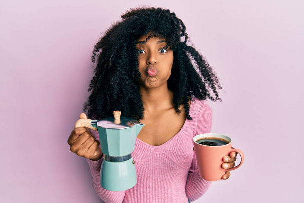 Mujer afroamericana con cabello afro bebiendo café italiano soplando mejillas con cara divertida. boca inflada con aire, captación de aire.  - Foto, imagen