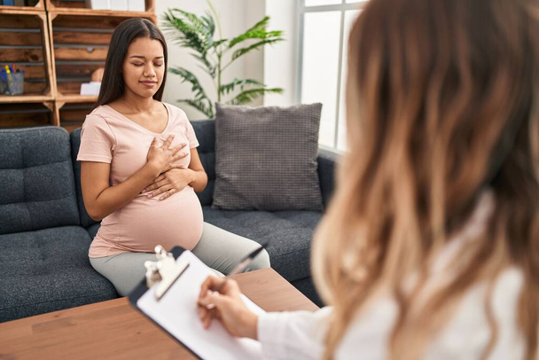 Jonge zwangere vrouw in therapie sessie glimlachend met handen op de borst met gesloten ogen en dankbaar gebaar op het gezicht. Gezondheidsconcept.  - Foto, afbeelding