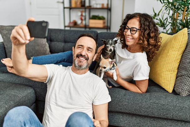 Ein hispanisches Paar mittleren Alters lächelt glücklich und macht Selfies mit dem Smartphone. Mit Hunden zu Hause auf dem Sofa sitzen. - Foto, Bild